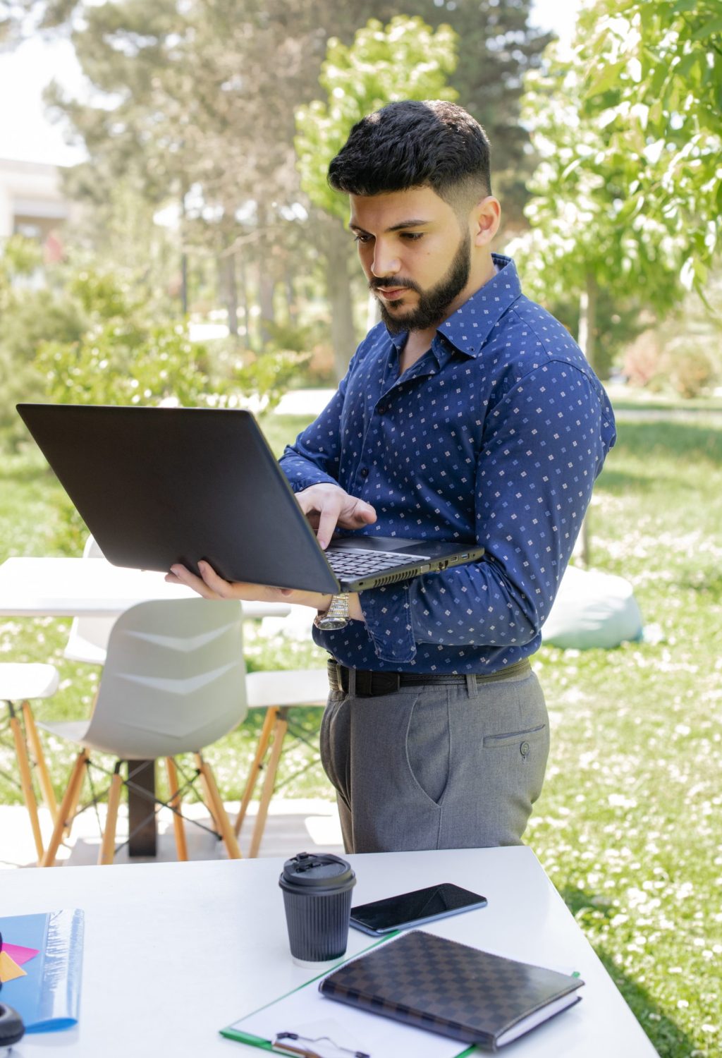 Handsome caucasian businessman typing on laptop outdoors. Portrait view of young black haired man in trendy shirt holding notebook, while working, with sunlit backyard on background. Concept of work.